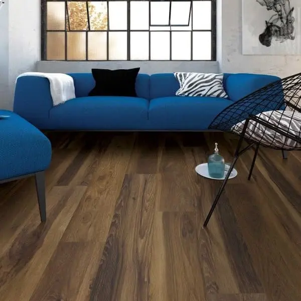 Waterproof vinyl planks in a modern living room with blue sectional sofa, black wire-framed chair, and industrial-style windows.