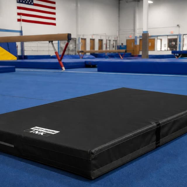 Black crash mat on blue gymnastics floor in gym, unfolded with straps and logo visible