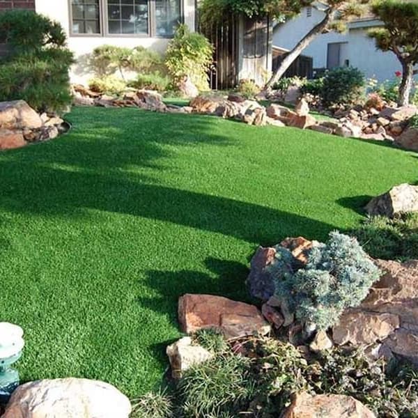 Artificial turf rolls in a residential backyard surrounded by rocks with a brick walkway.