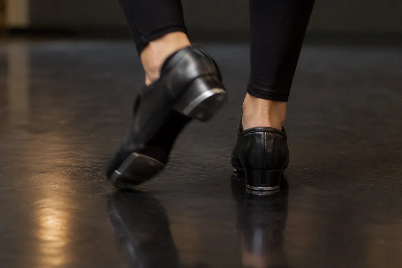 Close-up of black tap shoes mid-step on a glossy black marley dance floor