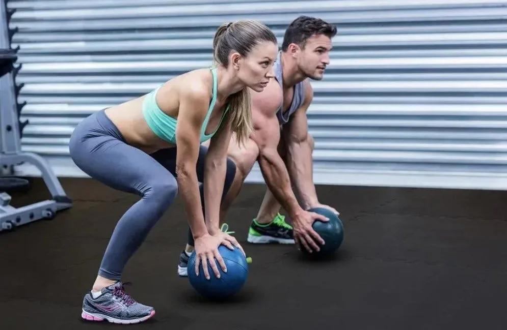 A man and woman perform squats while holding medicine balls on black interlocking rubber gym flooring. The durable gym mats provide a stable, non-slip surface that is ideal for dynamic workouts and functional training.