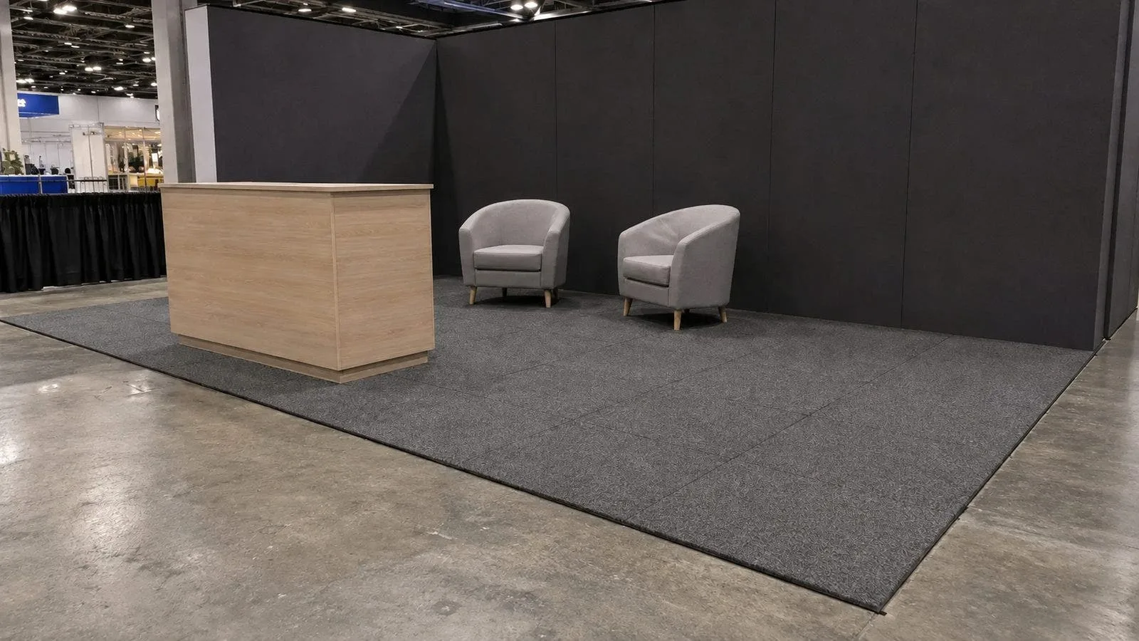 Trade show booth floored wall to wall with gray interlocking carpet tiles, reception counter and two chairs