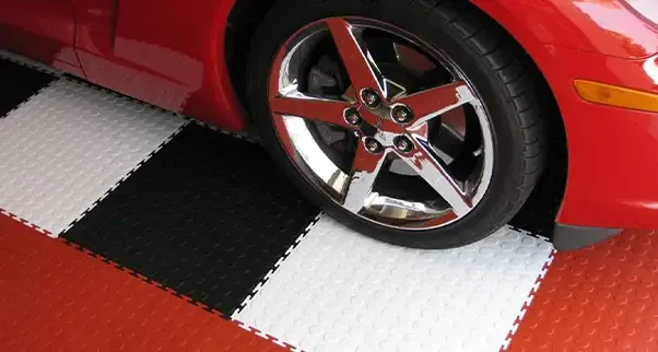 Checkerboard Coin-Top Garage Tiles The chrome wheel of a red sports car is parked on a checkerboard interlocking garage floor. The coin-top garage tiles are arranged in a custom pattern of red, black, and white, creating a durable and visually striking surface for vehicle display.