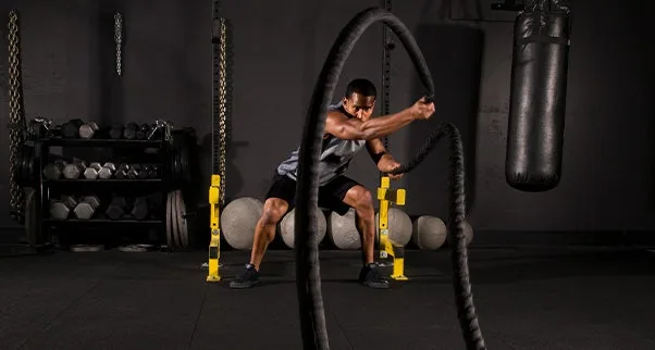 Rubber Flooring Banner An athlete performs a high-intensity battle rope workout on black rubber gym flooring. The durable commercial-grade floor tiles protect the subfloor and provide a stable surface for dynamic exercises and heavy equipment like dumbbells and a punching bag.