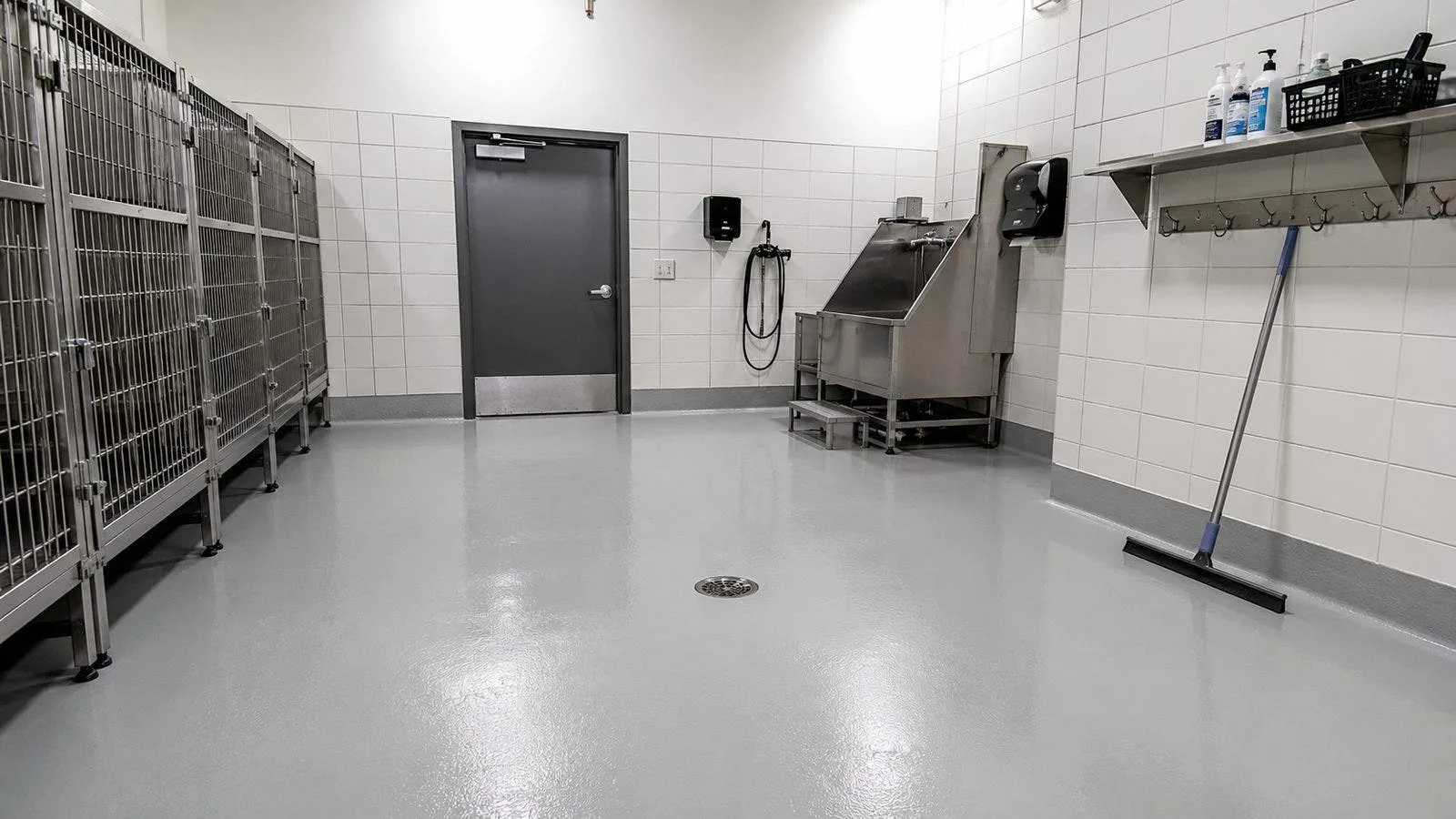 Kennel washdown and grooming room with a seamless coated floor sloping to a center drain, a stainless bathing tub, wall-mounted sprayer and a squeegee against the wall.