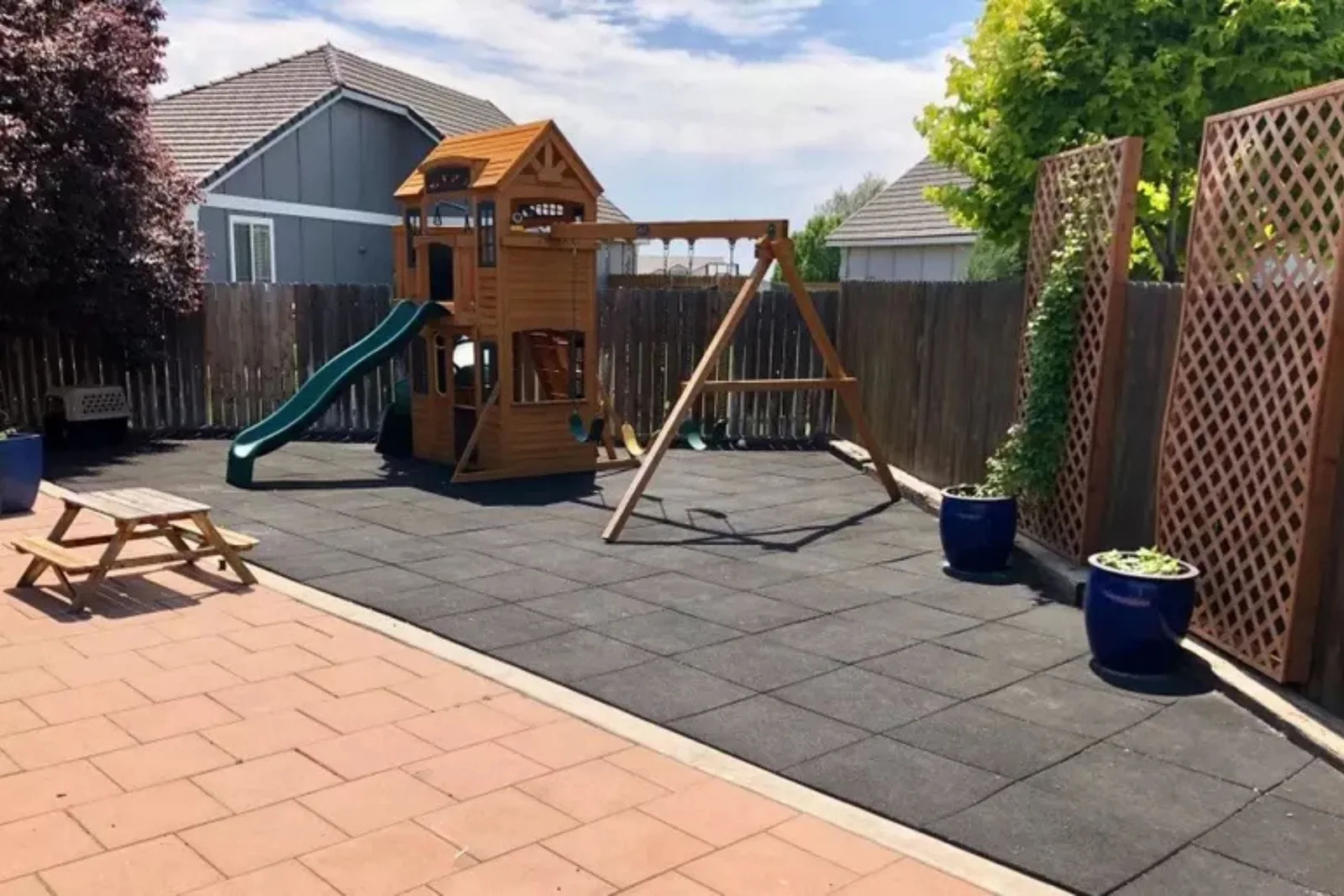 Durable black rubber playground tiles Backyard playground area with a wooden playset, swing set, and slide installed over durable black rubber playground tiles.