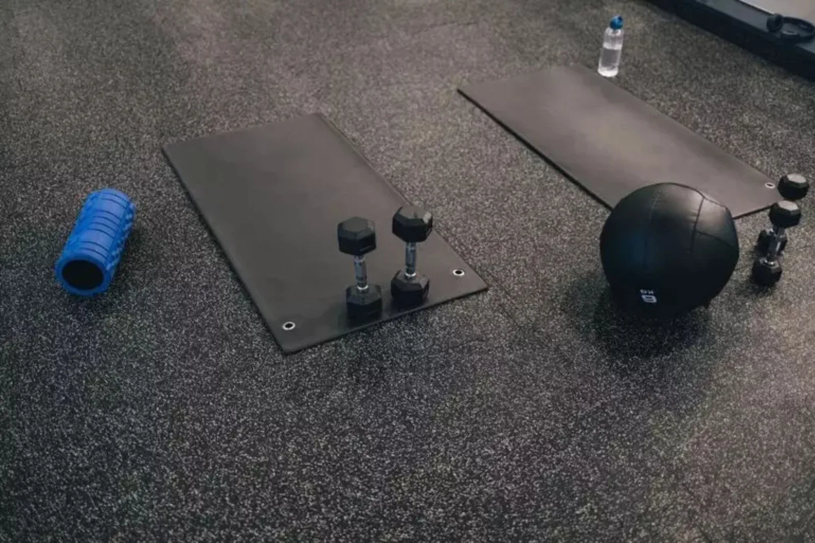 Durable black speckled rubber gym flooring Home gym setup with exercise mats, dumbbells, a medicine ball, and a foam roller on durable black speckled rubber gym flooring.