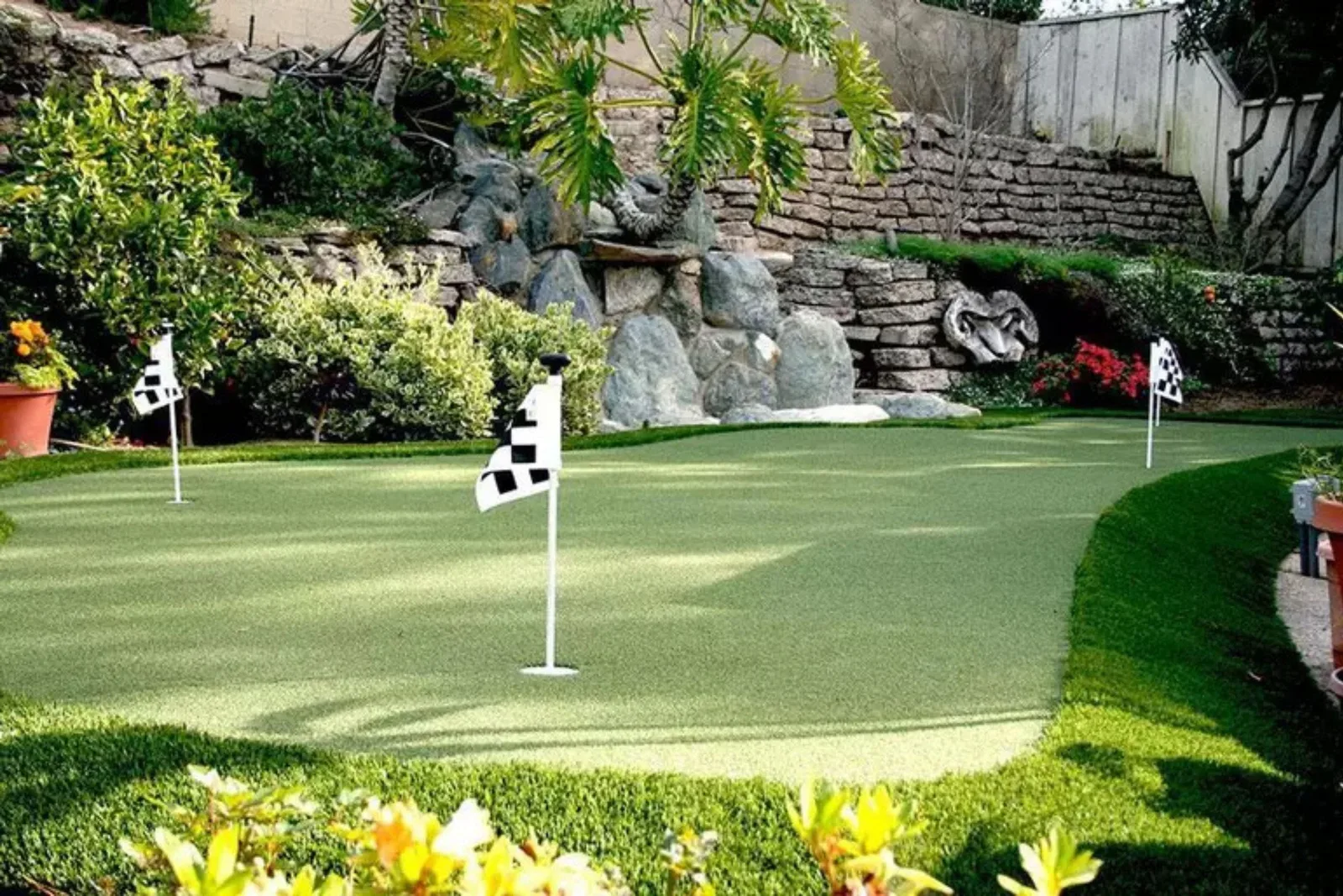 Synthetic Turf Putting Green Installation Professional backyard putting green with synthetic turf, stone retaining walls, and checkered golf flags in a lush garden setting.