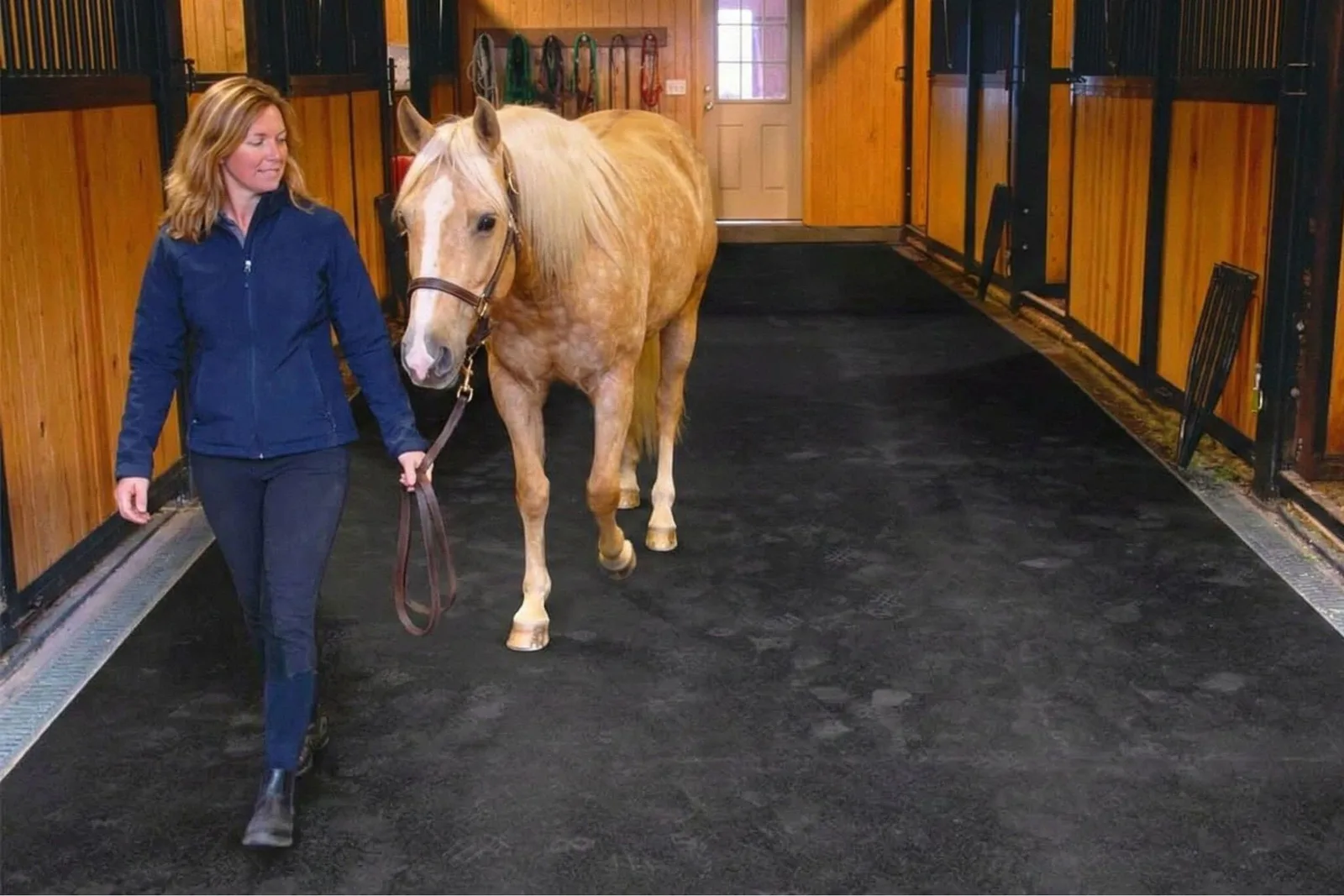 Woman leading a palomino horse down a barn aisle on rubber stall mats