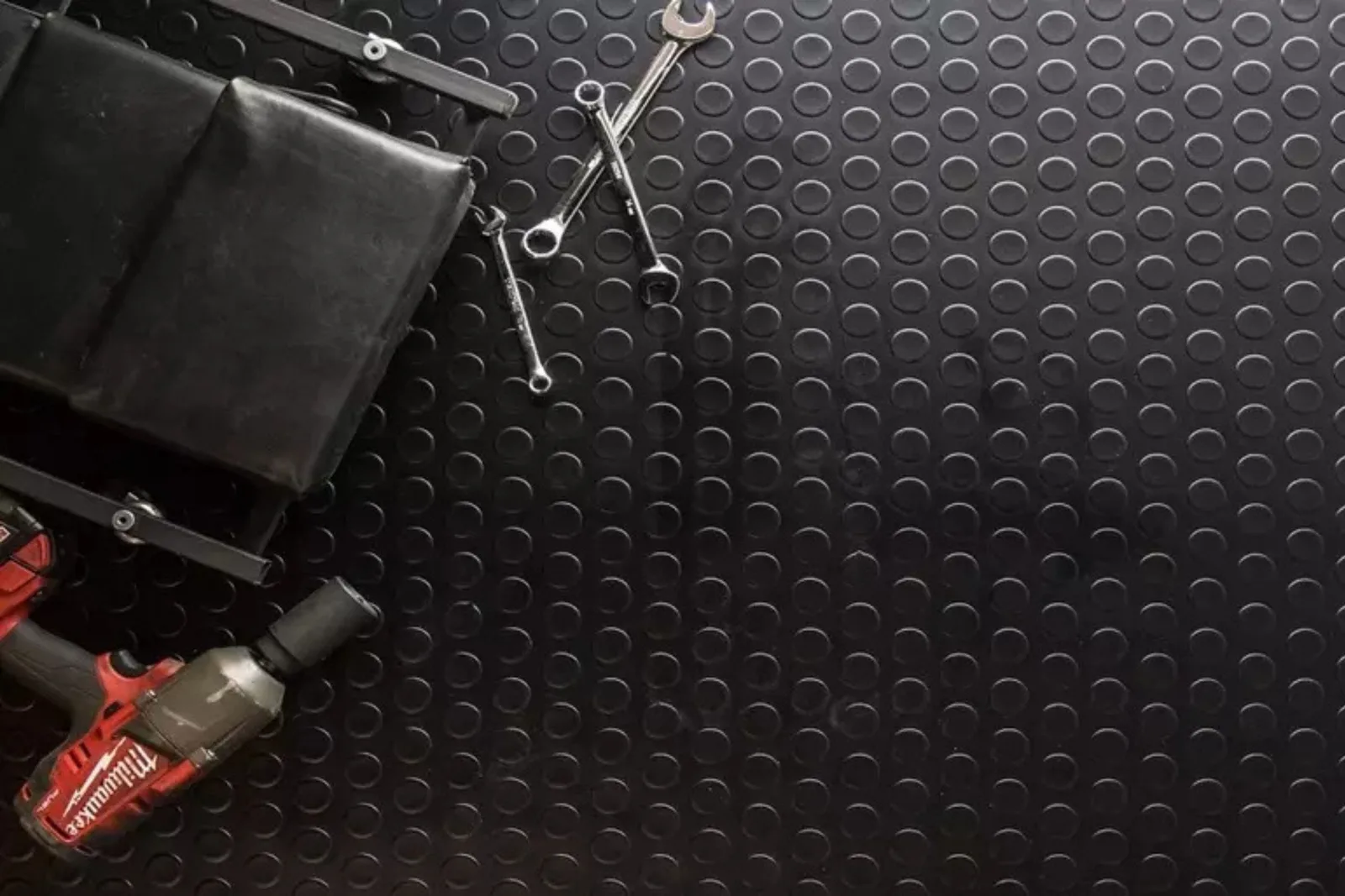 Coin-pattern black garage flooring roll with wrenches, a drill and a creeper seat