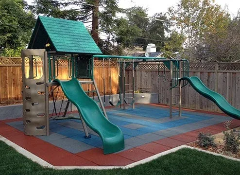 A backyard playground with a play structure, slides, and swings, installed on interlocking rubber playground tiles. The outdoor safety flooring features a colorful pattern of blue, red, and gray tiles, providing a shock-absorbent and protective surface fo