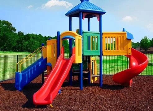 A colorful playground featuring a blue and yellow play structure, slides, and swings, all installed on a surface of vibrant red interlocking rubber safety tiles. The outdoor safety flooring provides a protective, shock-absorbent base for children's play a