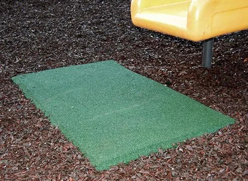 A green rubber playground swing mat placed at the bottom of a yellow playground slide, surrounded by brown rubber mulch. This durable playground safety mat is designed to withstand high-traffic use and reduce surfacing displacement in heavy-wear zones.