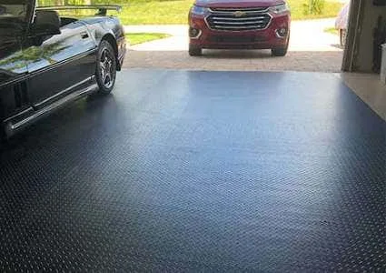 A garage floor covered entirely with seamless black diamond-plate garage flooring. A black sports car and a red SUV are partially visible, demonstrating how the durable roll-out garage matting protects the concrete and provides a clean, professiona