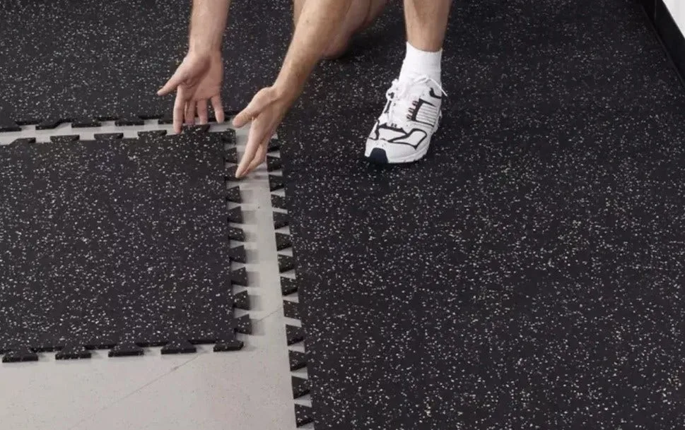 A person installing black interlocking rubber floor tiles with white speckles. This image demonstrates the simple assembly process of DIY gym flooring, showing the puzzle-piece edges connecting to create a seamless, professional-looking surface.