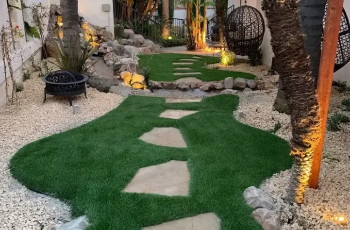Landscape turf rolls provide a low-maintenance and lush green artificial grass landscaping solution, with a curving stone pathway and a fire pit in a professionally designed backyard