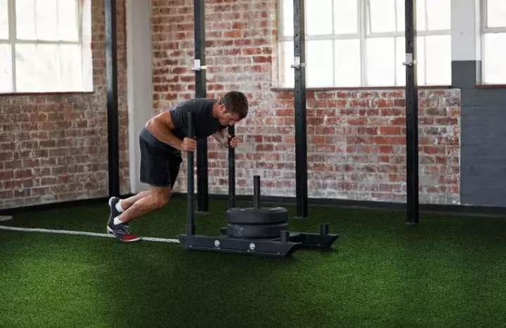 Artificial grass gym turf provides a durable and low-impact flooring solution for athletic training, with a man performing sled pushes on the green turf in a brick-walled gym.