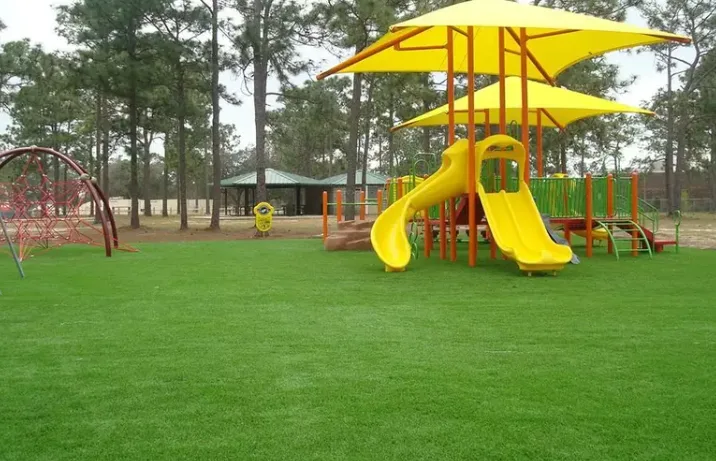Playground turf provides a safe, low-maintenance, and lush green artificial grass surface for a public park playground, featuring a yellow slide and a colorful play structure.