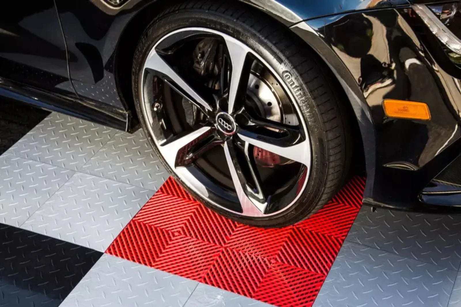 The wheel of a black Audi sports car is parked on a section of interlocking garage flooring tiles. The floor features a checkerboard pattern of gray, white, and red, highlighting the customizable and durable surface for automotive use.
