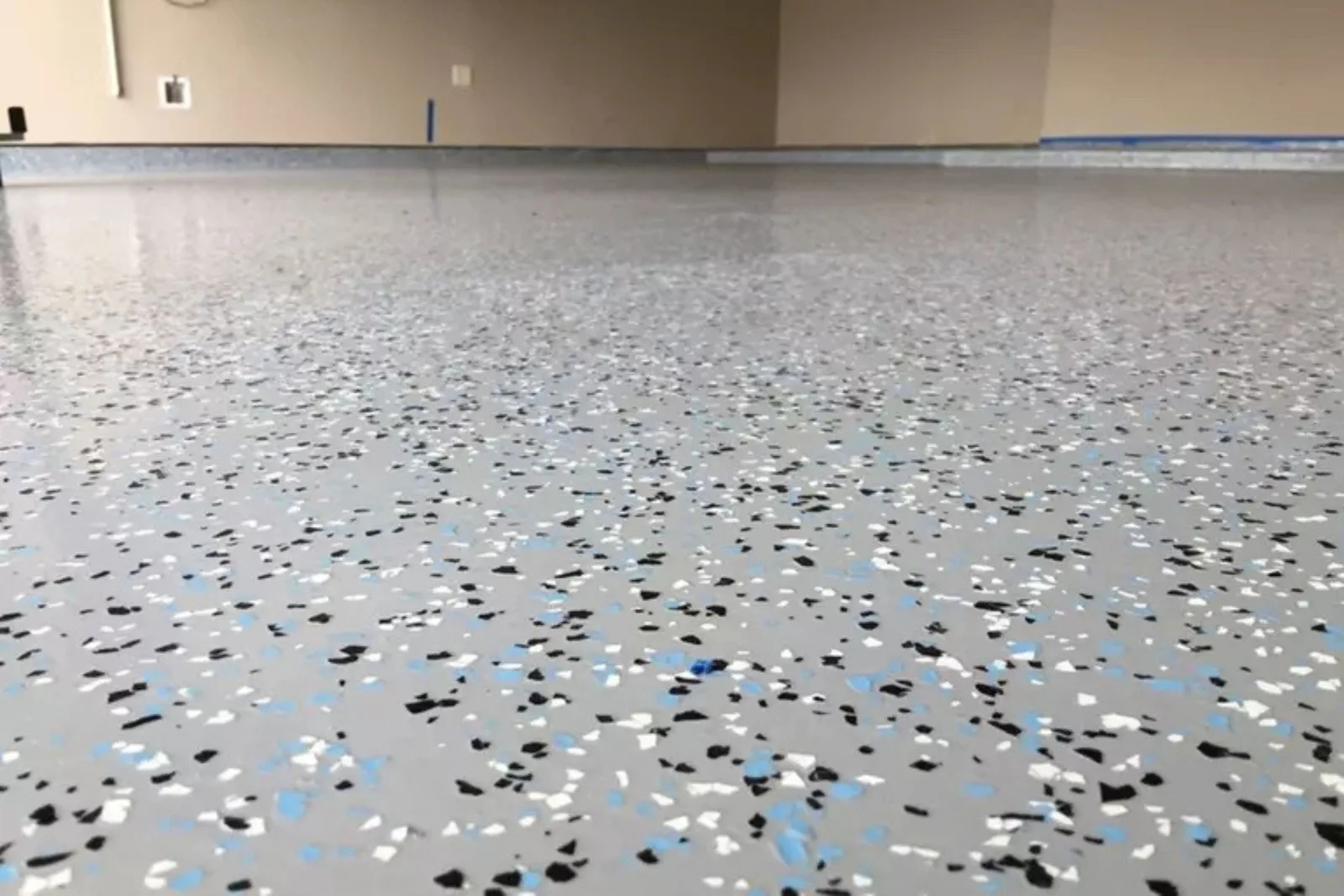 A close-up view of a newly installed epoxy garage floor with a smooth, glossy finish. The light gray surface is covered in decorative flakes of black, white, and light blue, providing a durable and attractive surface for a garage.