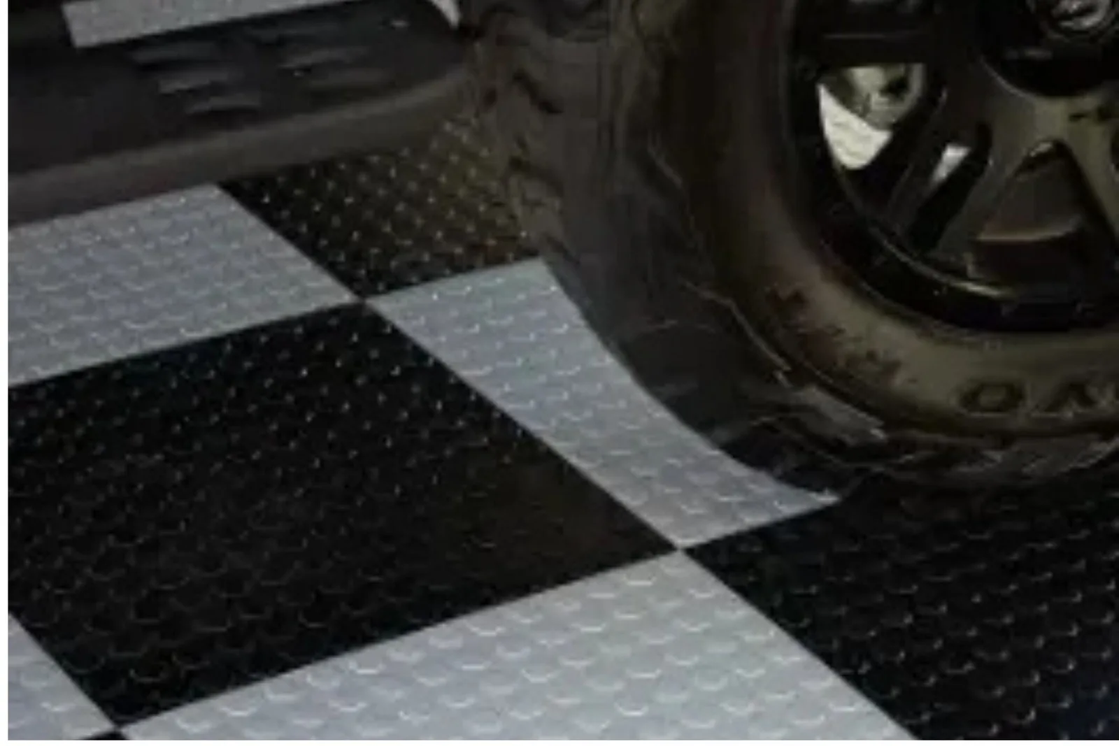 A close-up of a vehicle tire on a checkerboard garage floor made of black and gray coin-top interlocking tiles. This image highlights the textured, non-slip surface of the durable flooring, which is ideal for a garage or workshop.