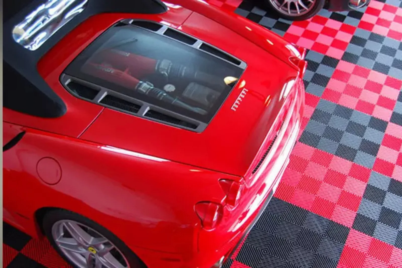 An overhead view of a sleek red Ferrari convertible parked on a custom interlocking garage floor. The floor features a high-end checkerboard design of red, gray, and black garage floor tiles, providing a striking and protective surface for luxury vehicles