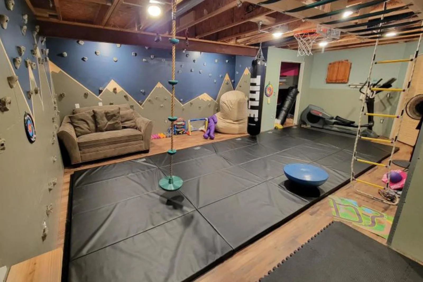 Folding foam panel mats in black create a large, cushioned, and safe home gym flooring area for acrobatics, exercise, and climbing wall use in a basement.