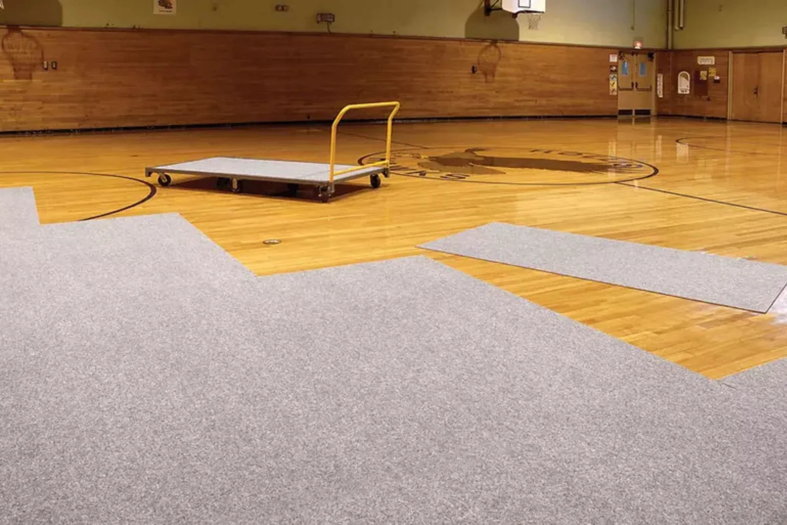 Gym floor cover carpet tiles in light gray are laid out on a polished hardwood basketball court, demonstrating a portable gymnasium flooring protection solution for special events.
