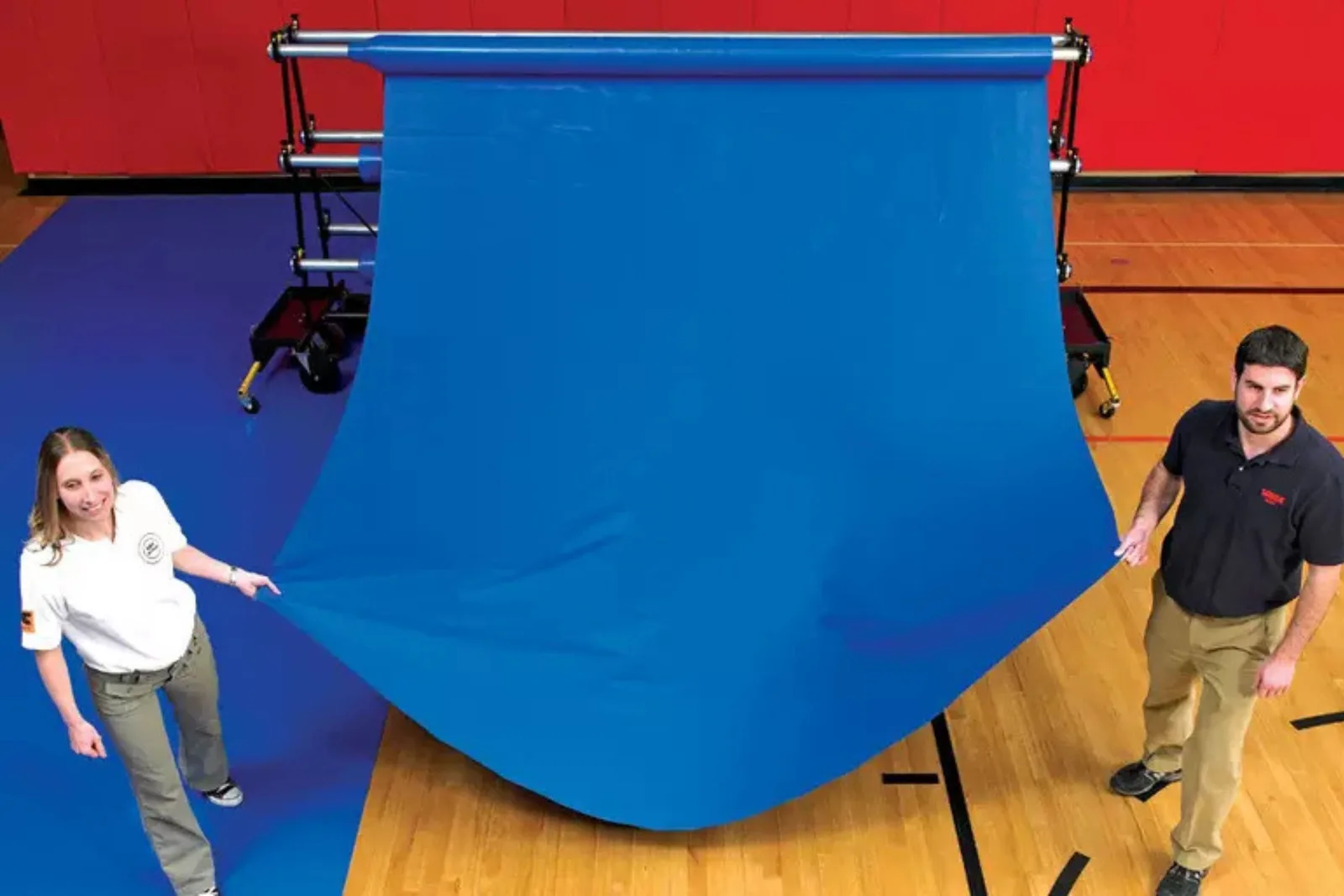 Rolled vinyl gym floor covers in blue are unwound from a mobile storage rack, demonstrating a portable and easy-to-deploy gymnasium floor protection solution for events.