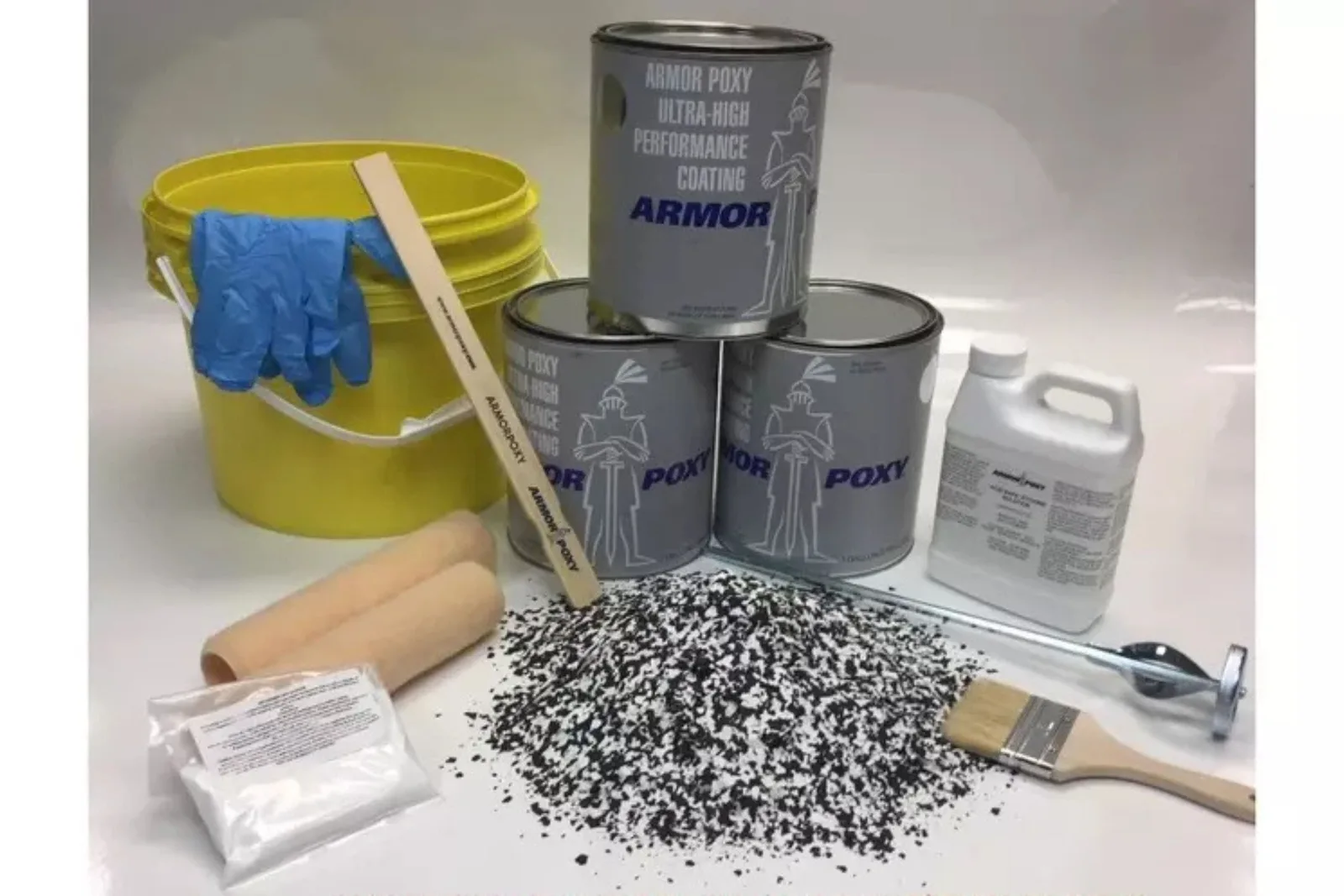 A complete DIY epoxy floor coating kit laid out for application, including three cans of epoxy resin, a yellow mixing bucket, rollers, a stir stick, a bag of anti-slip powder, a paint brush, and a large pile of black and white decorative floor flakes.