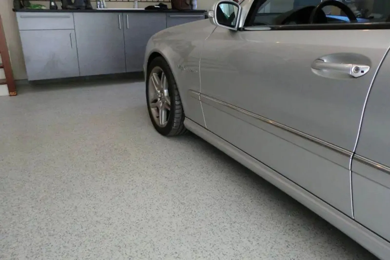  A silver convertible is parked in a clean garage with a light gray epoxy floor coating. The durable decorative flake garage floor provides a seamless, high-gloss, and stain-resistant finish that complements the modern metal cabinets.