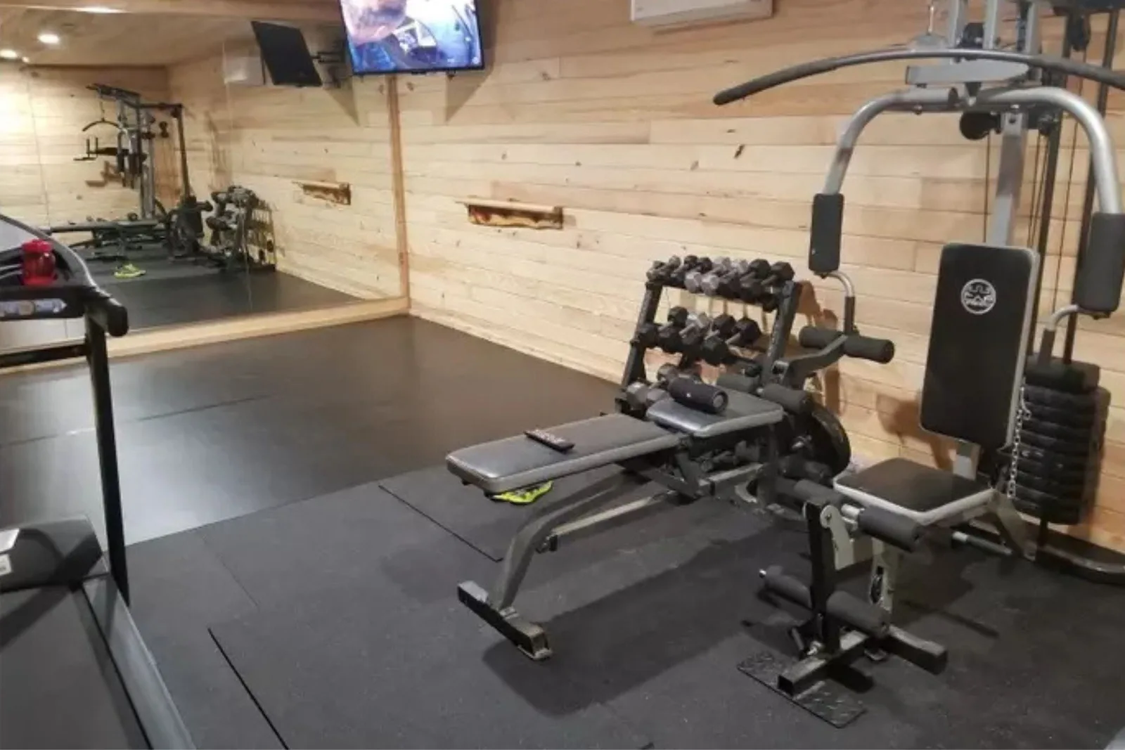 A cozy home gym with light wood-paneled walls and a large mirror, featuring black rubber gym flooring. The seamless rubber mats provide a protective, durable, and stable base for a weight machine, dumbbell rack, and a treadmill.