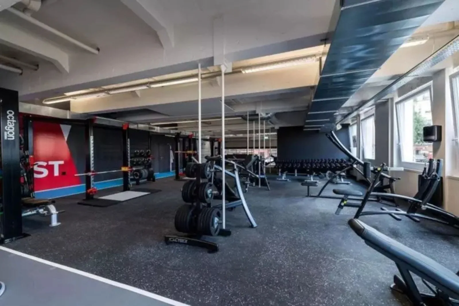 An industrial-style commercial gym with a large, continuous installation of black speckled rubber gym flooring. The heavy-duty rubber floor provides a protective and stable surface for numerous free weights, squat racks, benches, and other fitness equipme