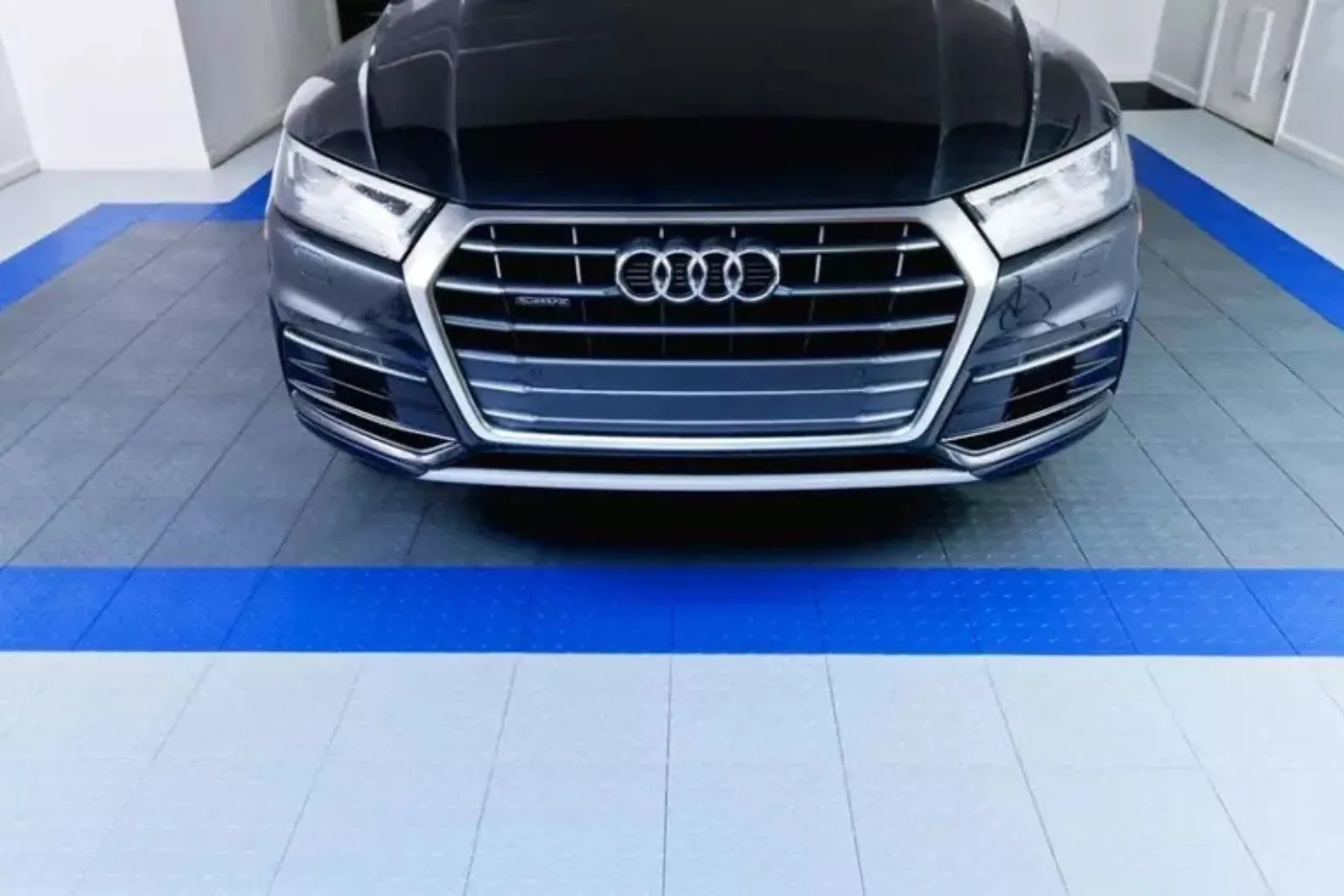 The front of a black Audi SUV is parked on interlocking garage flooring tiles in a custom design. The floor features white modular tiles bordered by a striking blue racing stripe, providing a clean, durable, and high-end look for an automotive garage.