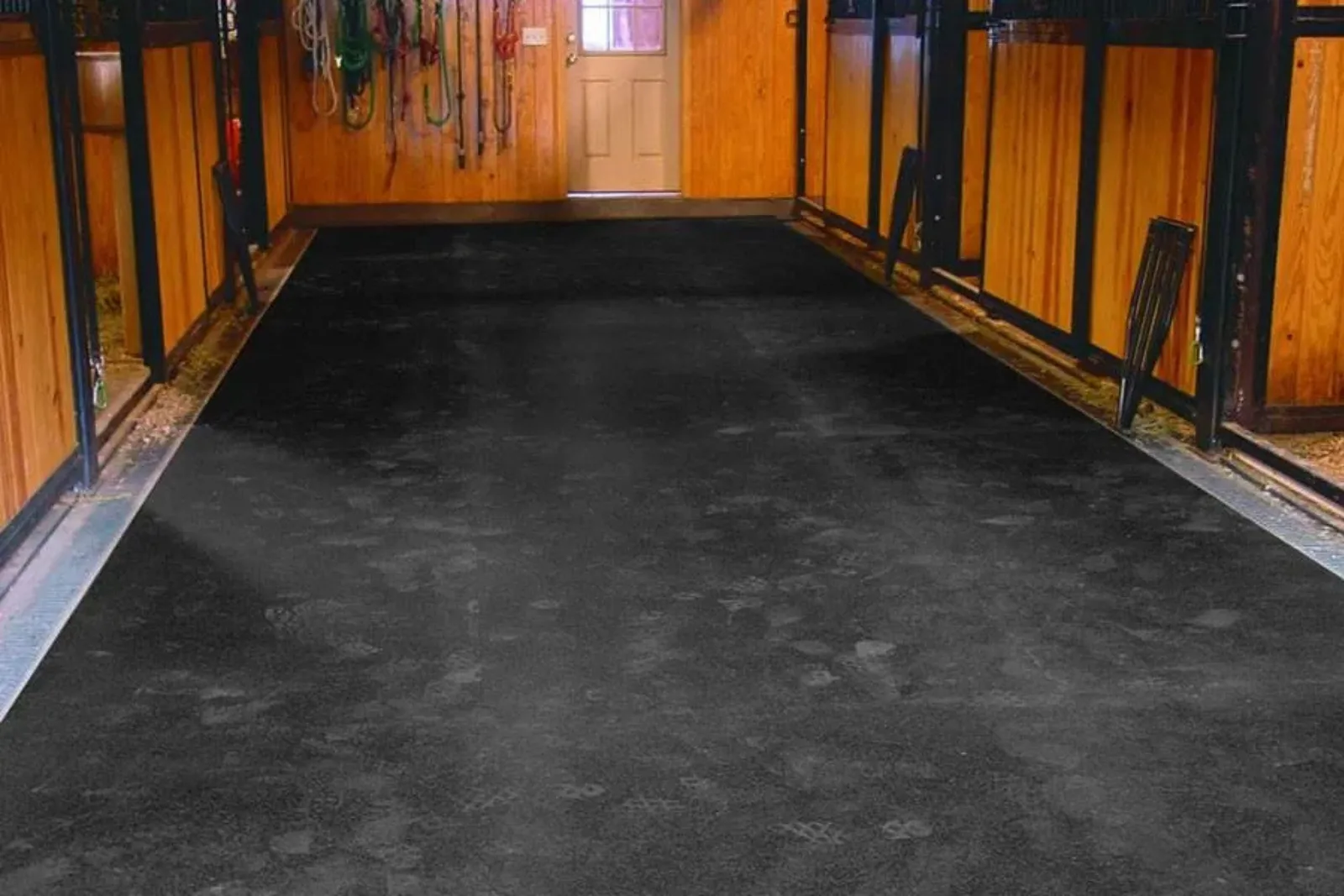 A wide aisle in a horse stable or barn, completely covered with seamless black rubber horse stall mats. This heavy-duty rubber flooring provides a non-slip, shock-absorbent, and hygienic surface for horses and stable workers, protecting the concrete subfl