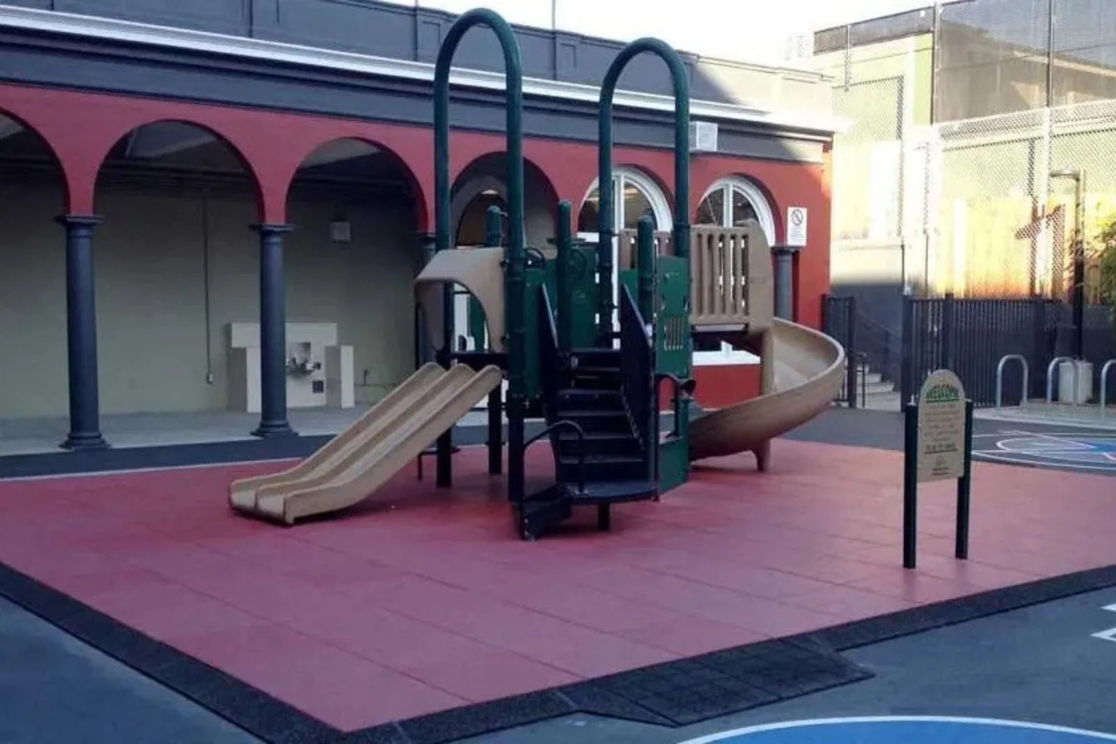 A playground featuring a climbing structure and slides, all placed on red interlocking rubber safety tiles. The durable playground flooring has a ramp for accessibility and provides critical fall protection for a clean, permanent play area near a school b