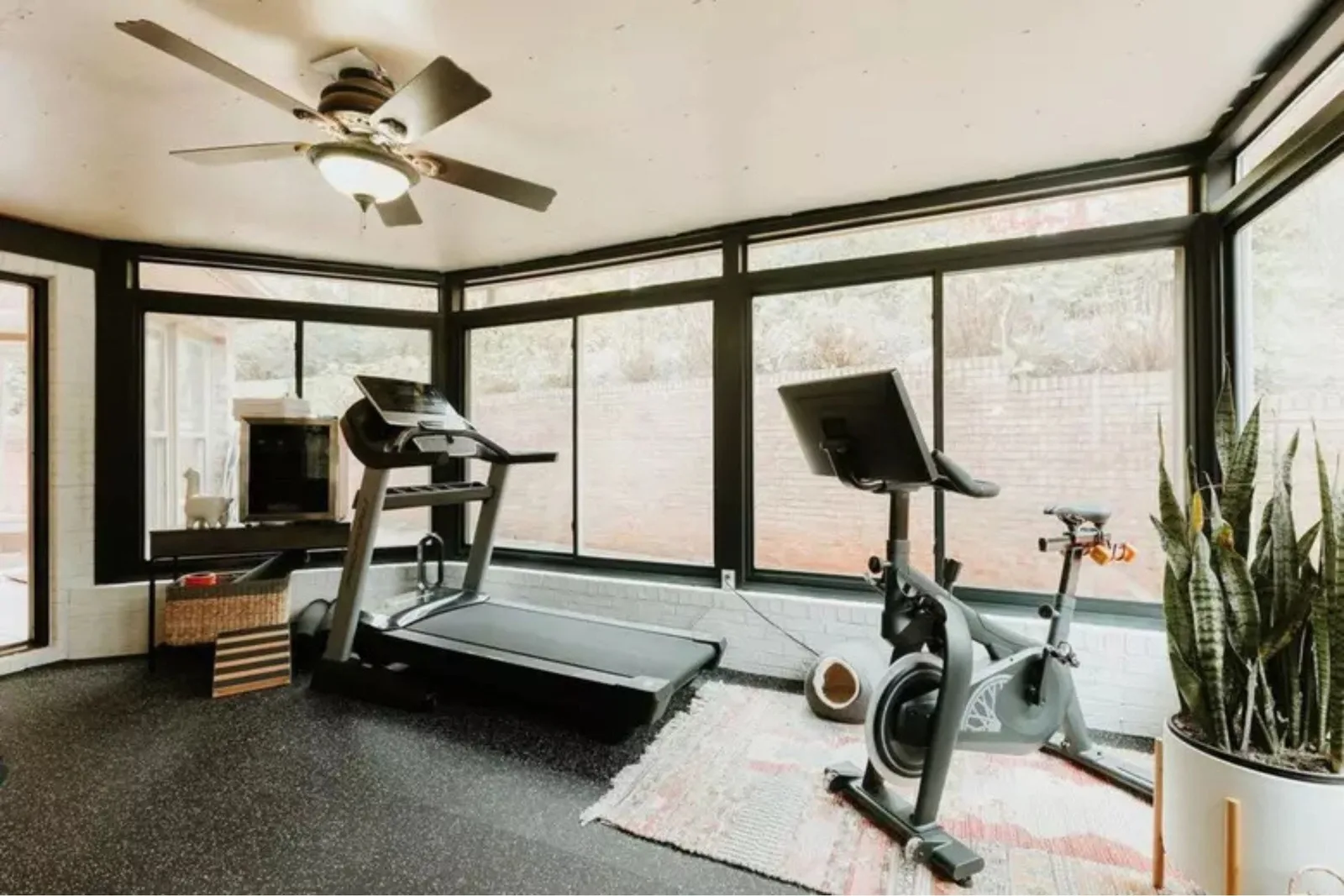 A bright home gym located in a sunroom, featuring a floor entirely covered in black speckled rubber gym flooring. The durable rubber matting provides a stable, protective surface for the treadmill and stationary bike, making it ideal for a home workouts