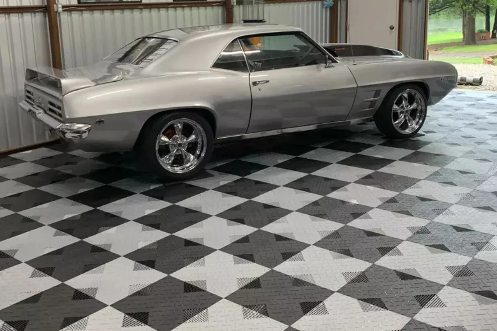 A classic silver muscle car is parked in a garage on a black, dark gray, and light gray checkerboard interlocking garage floor. The durable modular garage tiles create a high-end, protective, and custom display surface for the vehicle.
