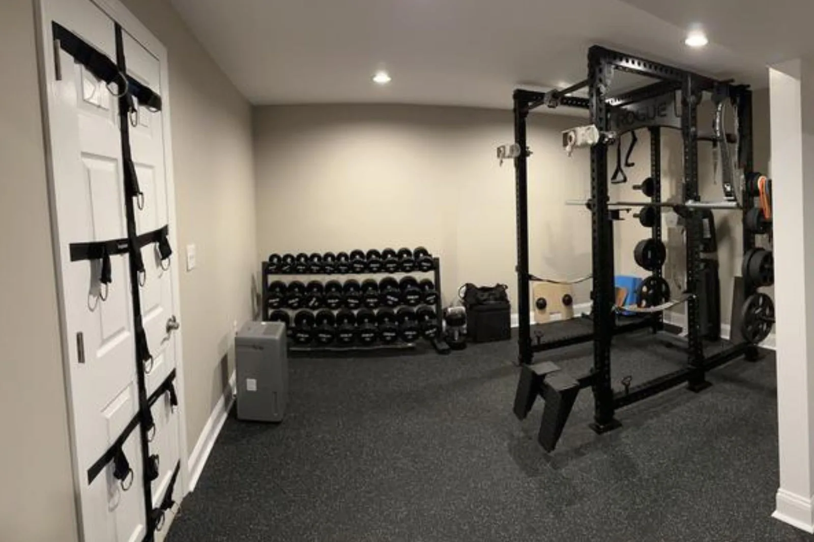 A well-organized home gym with black rubber gym flooring. The durable gym mats provide a protective and stable surface for a power rack, a full set of dumbbells on a storage rack, and various other workout accessories.