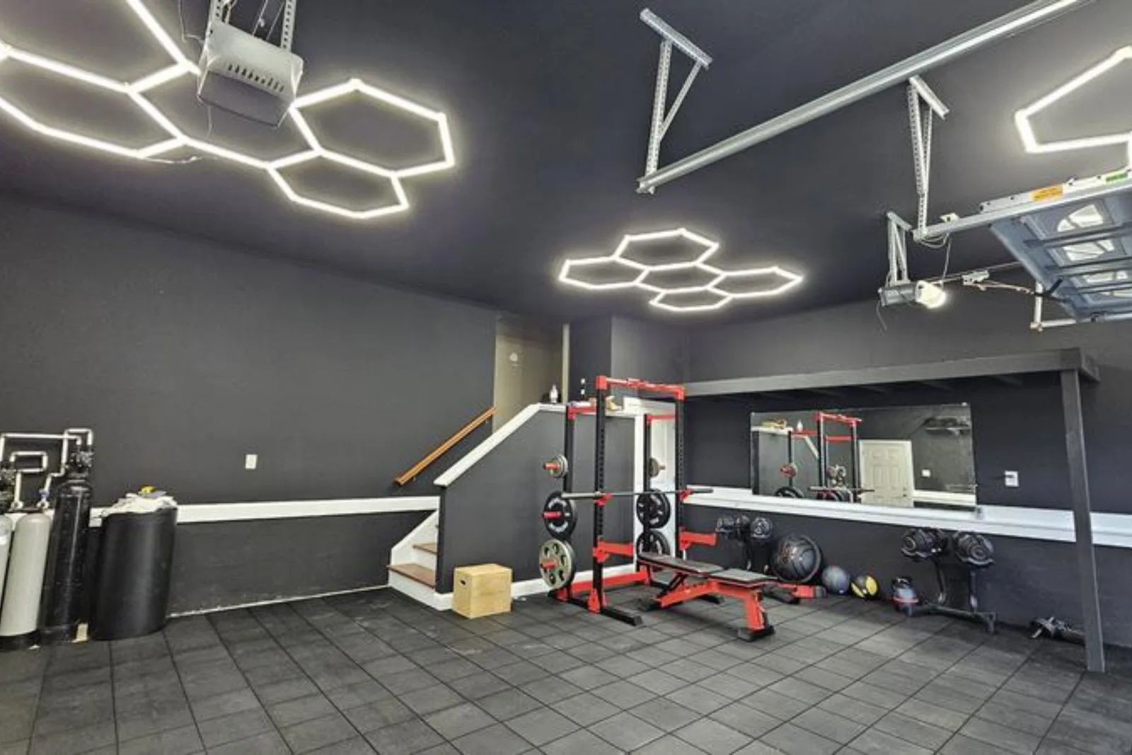 A modern garage gym with a dark aesthetic, featuring a floor of black interlocking rubber tiles. The durable garage flooring protects the concrete from heavy equipment like a power rack, weight bench, and dumbbells, all lit by unique hexagonal ceiling li