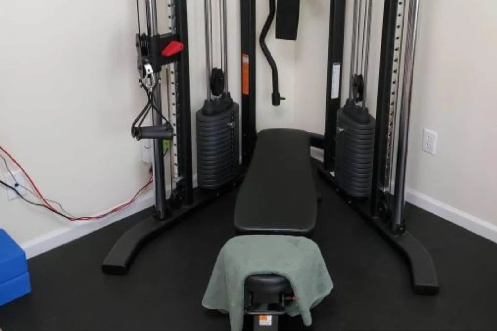 A compact home gym setup in the corner of a room, featuring black rubber gym flooring. The durable rubber mat provides a protective and stable base for a cable machine and weight bench, making the most of a small space.