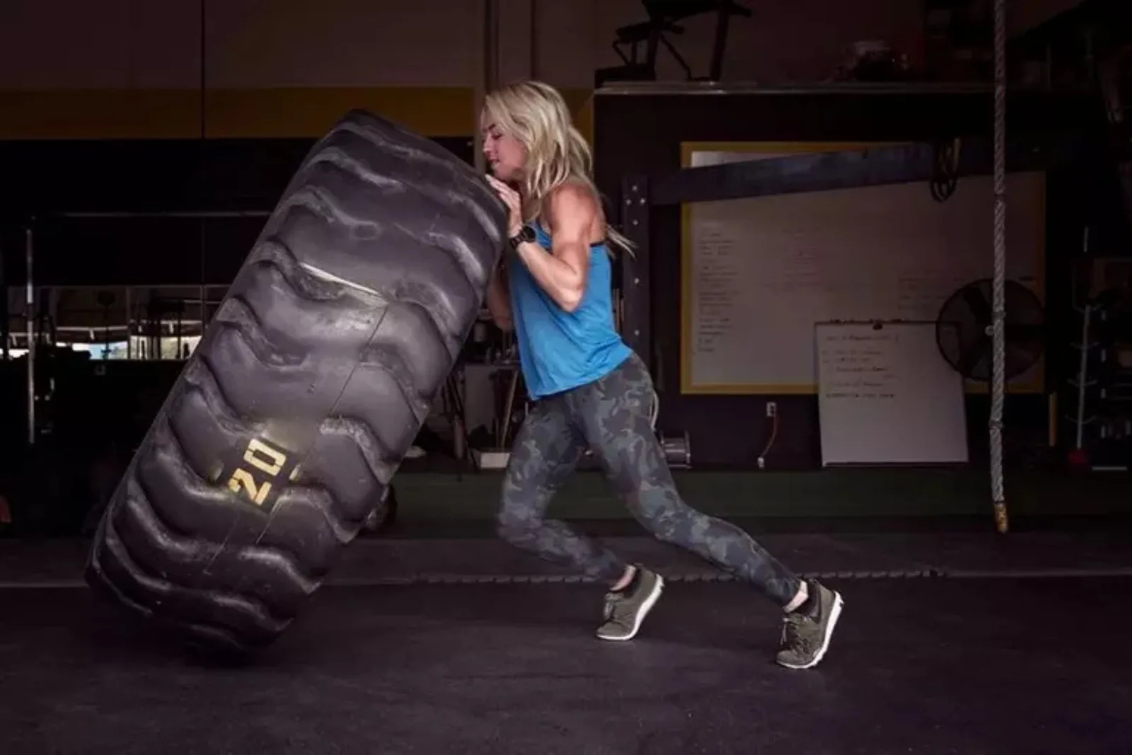 Heavy-duty rubber gym flooring is shown with a woman performing a tire flip in a workout facility. The durable rubber floor tiles provide a protective and stable surface, designed to withstand high-impact and heavy weight training.