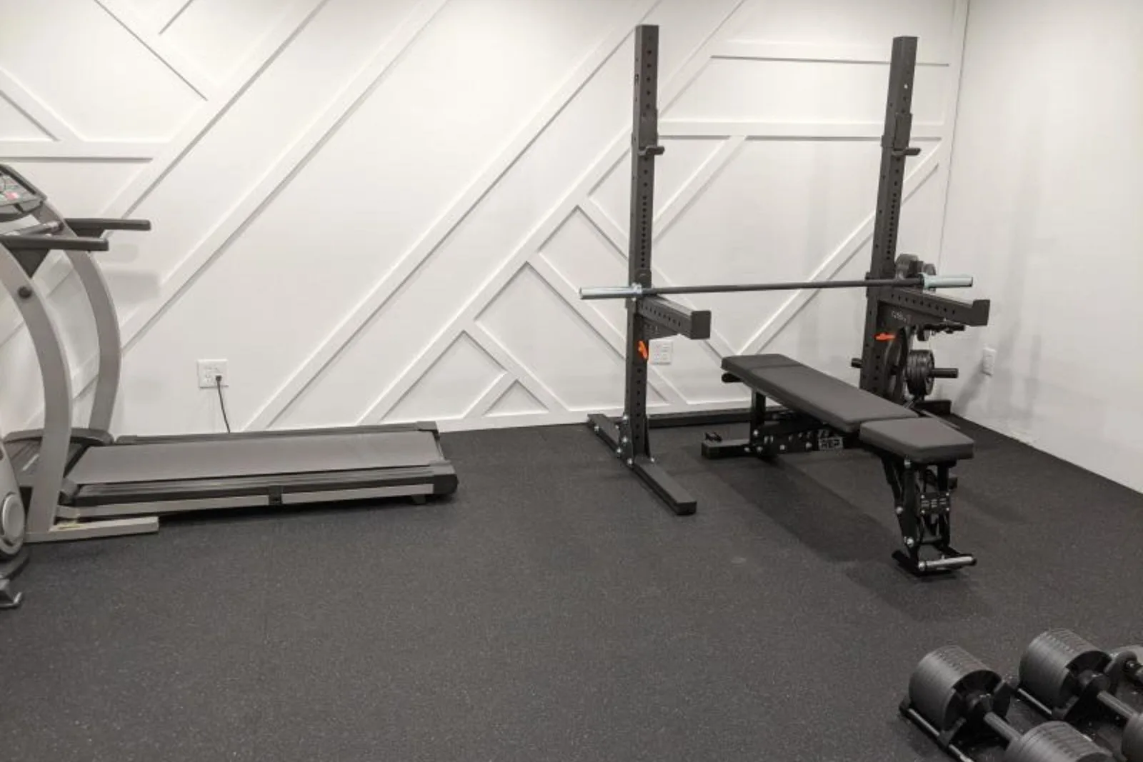A modern home gym with a black rubber gym floor providing a clean and protective surface for a treadmill and a squat rack with a weight bench. The durable rubber matting shields the subfloor from equipment and weight drops while creating a dedicated work