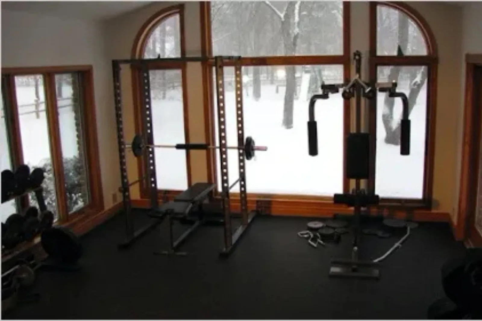 A home gym with a scenic window view of a snowy landscape, featuring black rubber gym flooring. The durable floor matting provides a solid foundation for a power rack, weight bench, and pec deck machine, protecting the subfloor from equipment and weight d