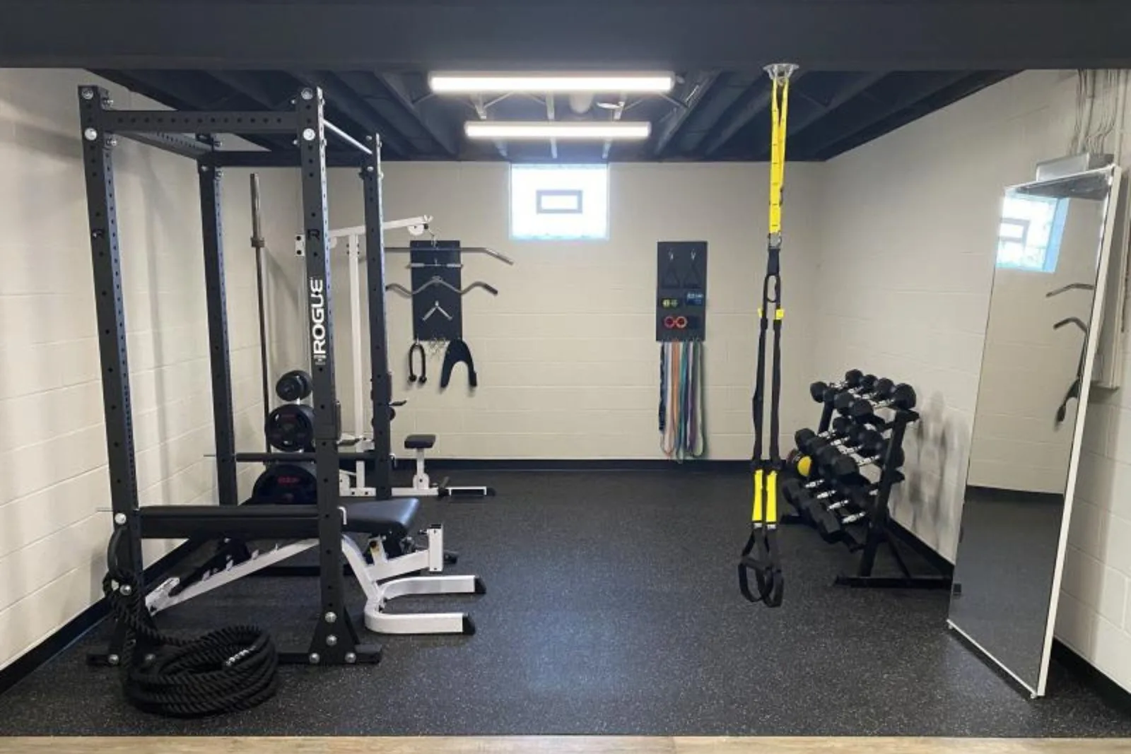 A complete home gym setup in a basement, featuring black speckled rubber flooring. The durable gym mats provide a protective surface for a power rack, bench, dumbbells, and resistance bands, creating an ideal and safe space for a variety of workouts.