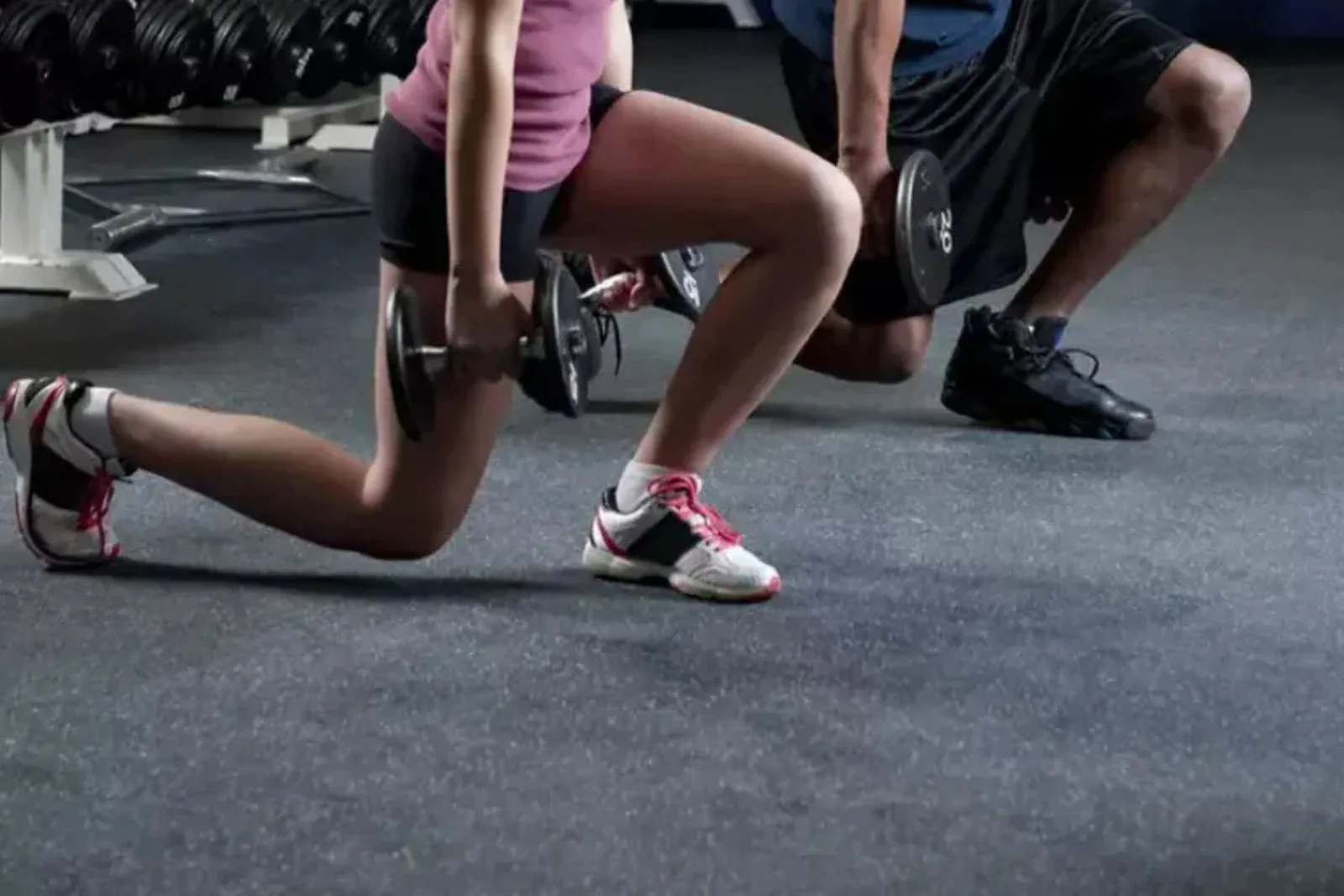 A man and woman perform dumbbell lunges on black speckled rubber gym flooring. The durable gym mats provide a non-slip, shock-absorbent surface for a variety of exercises, protecting both the subfloor and the joints of the users.