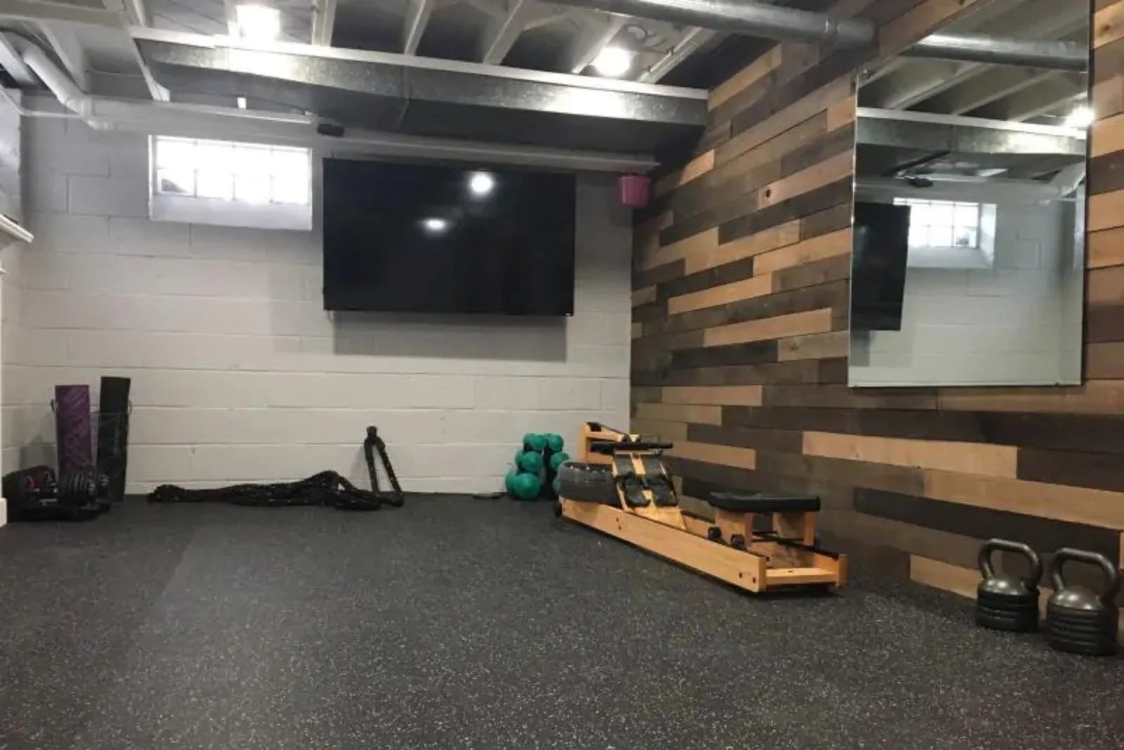A stylish basement home gym with black speckled rubber flooring. The durable gym matting provides a stable surface for a variety of equipment, including a rowing machine, dumbbells, kettlebells, and battle ropes, complemented by a wood-paneled accent wall