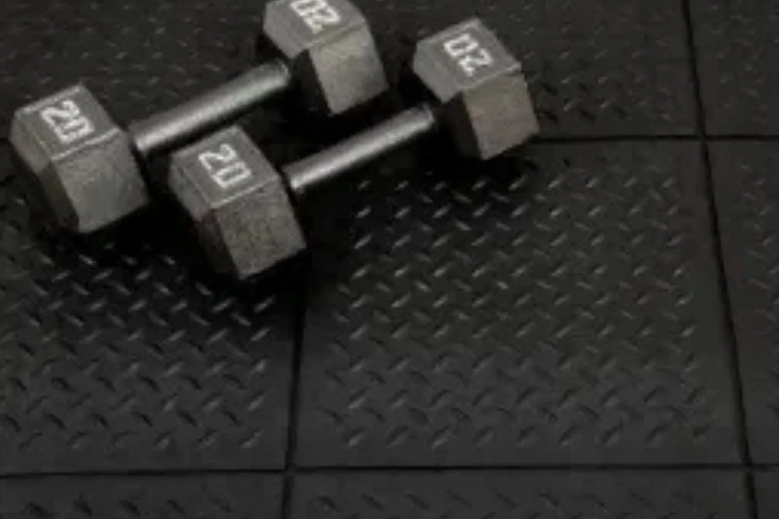 Dumbbells resting on a floor of black rubber gym tiles with a diamond plate texture. The interlocking rubber flooring provides a durable and protective surface for weightlifting and fitness activities.