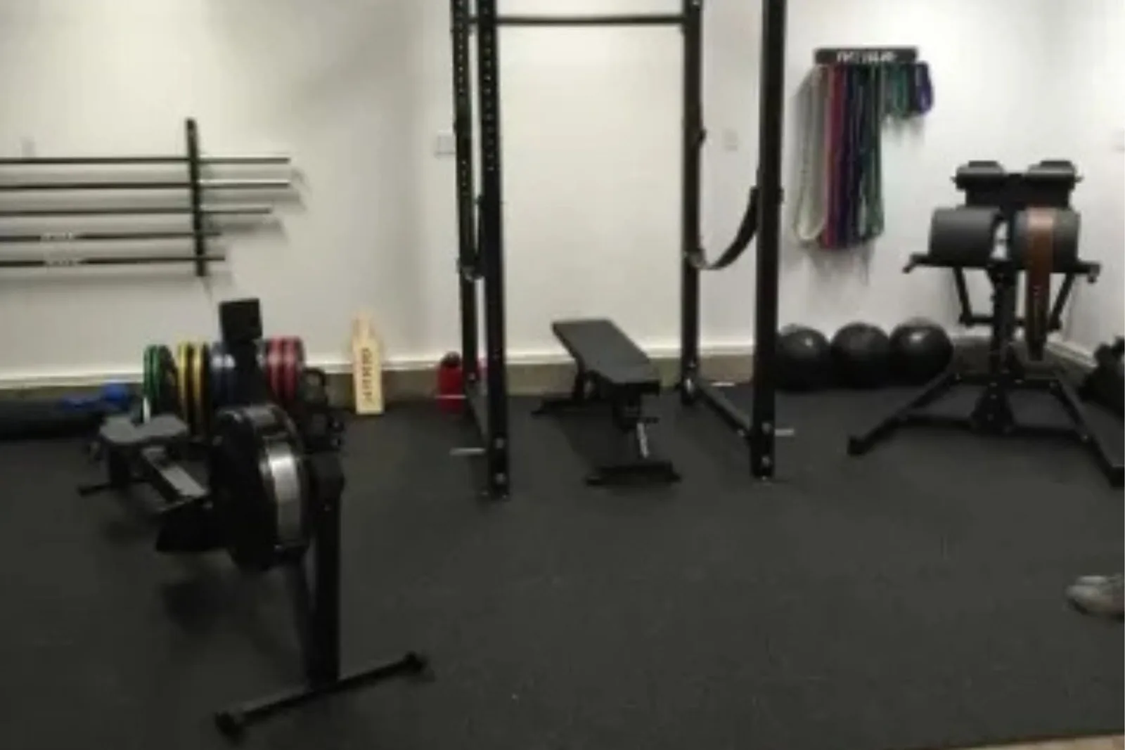 A clean, well-equipped home gym with a black rubber gym floor. The floor provides a protective and durable base for a squat rack, bench, rowing machine, and various weights and fitness accessories. The seamless rubber matting covers the entire workout are