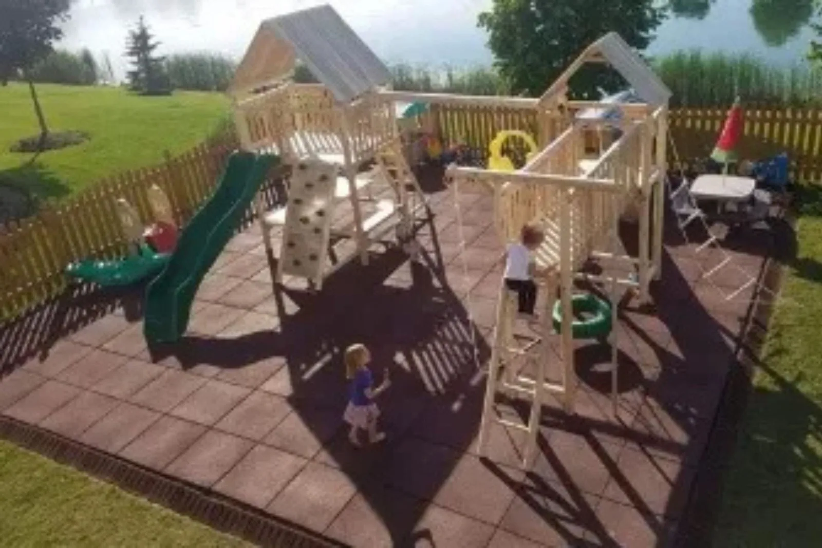 An outdoor children's playset with multiple slides and a climbing wall, situated on red rubber playground tiles. The durable outdoor matting creates a safe and soft landing surface, surrounded by a wooden fence and a grassy yard with a pond in the backgr